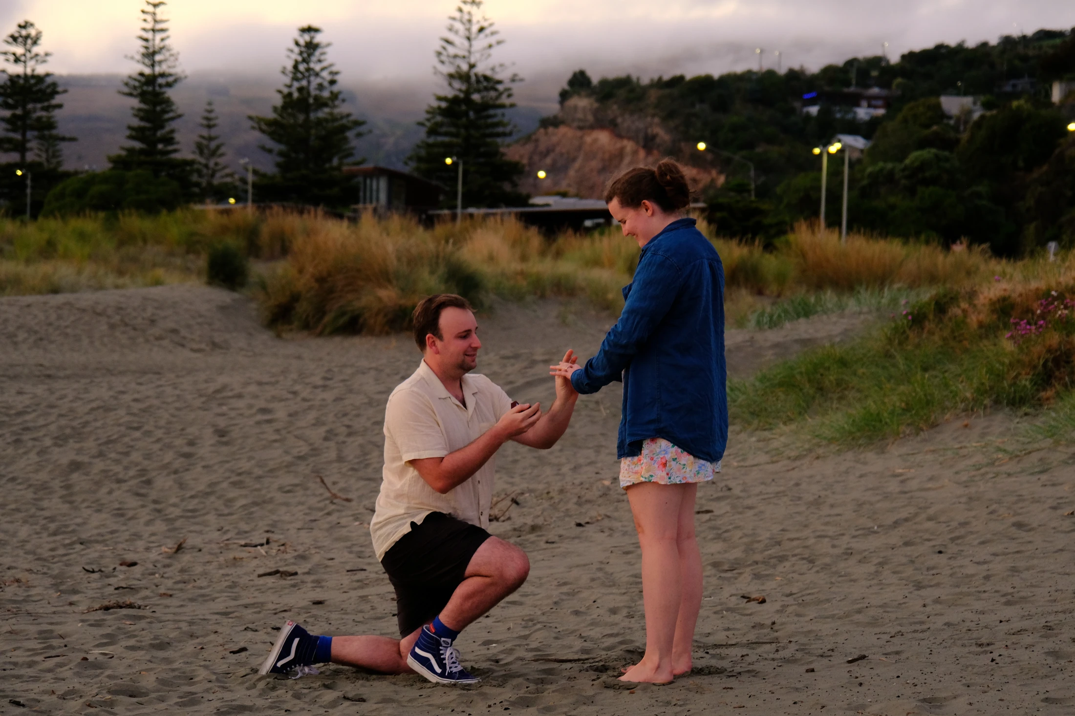 A man kneels to propose to his partner on a beach at dusk