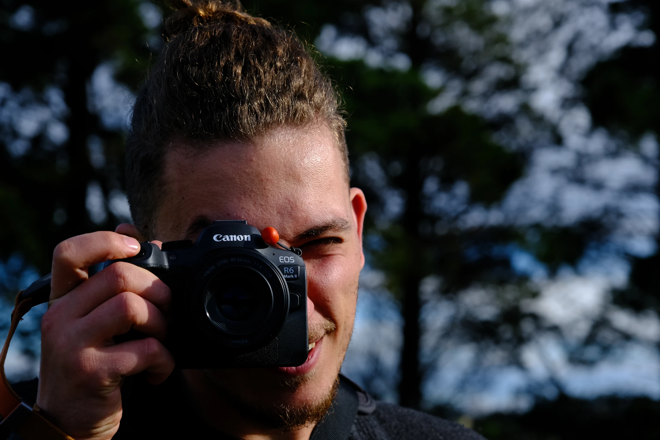 A photographer smiles from behind a Canon camera in dappled outdoor light