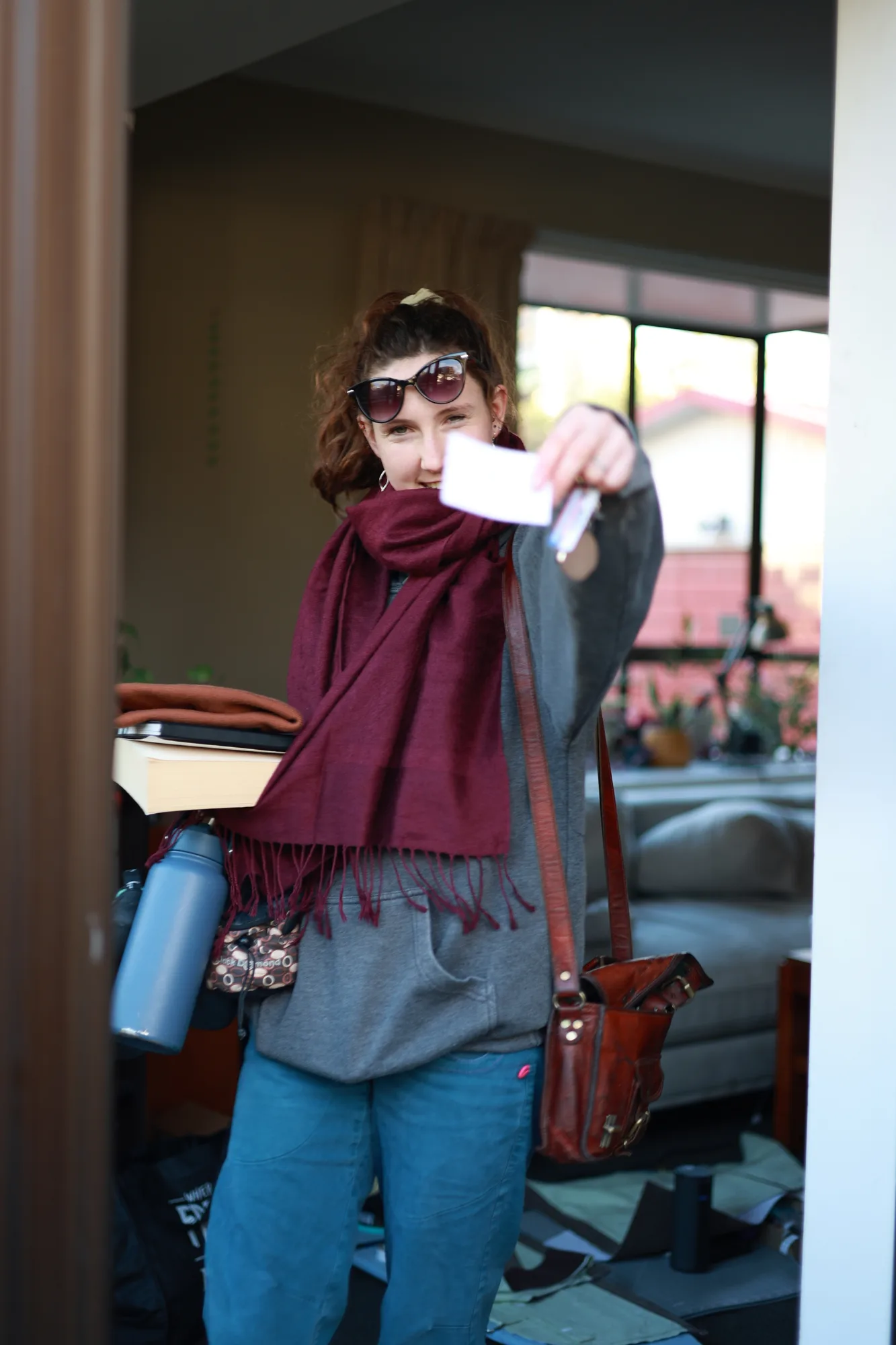 Caitlin stands in a doorway carrying books and bags while holding something towards the camera