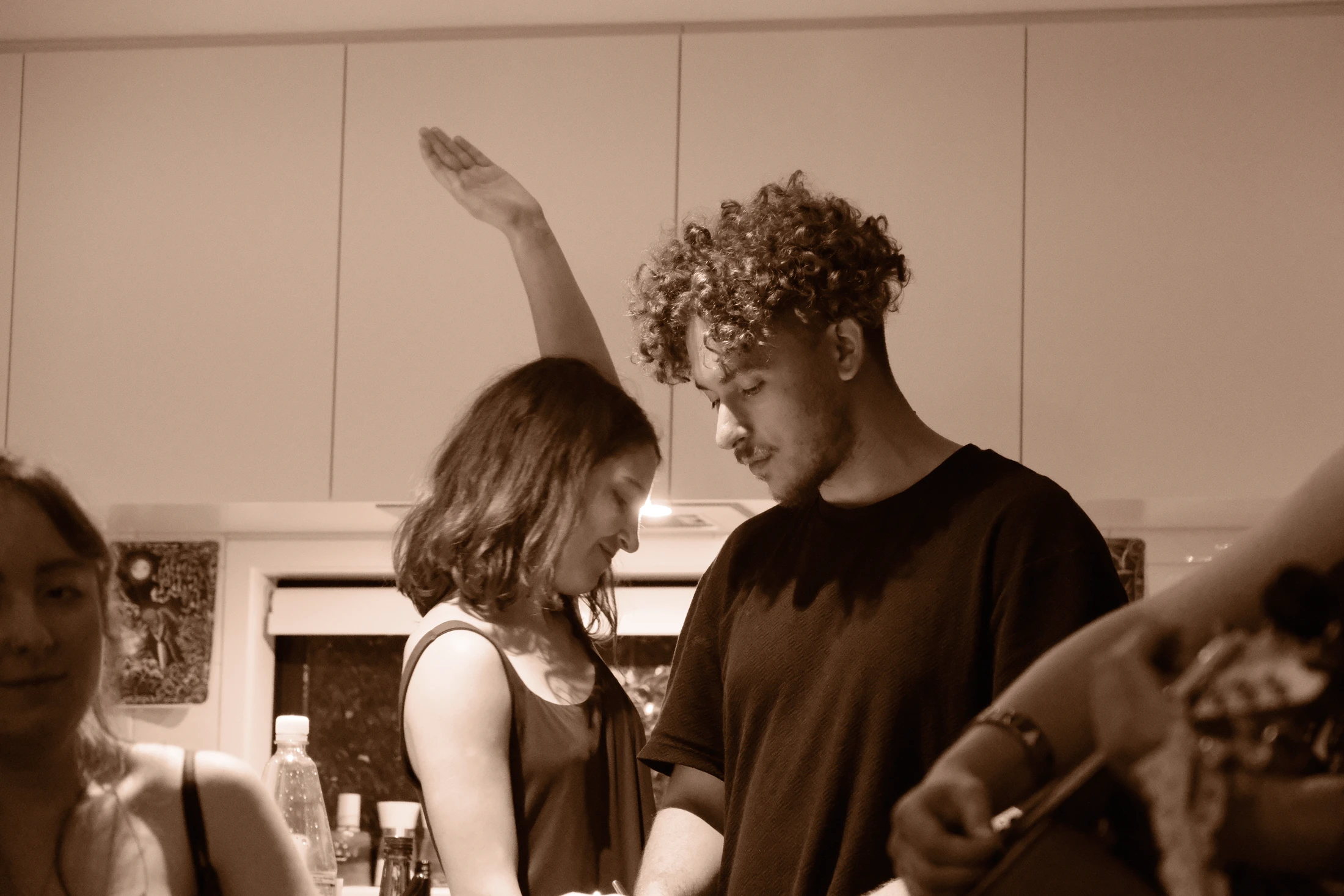 Two people lean close together while a raised arm crosses a warm sepia-toned scene