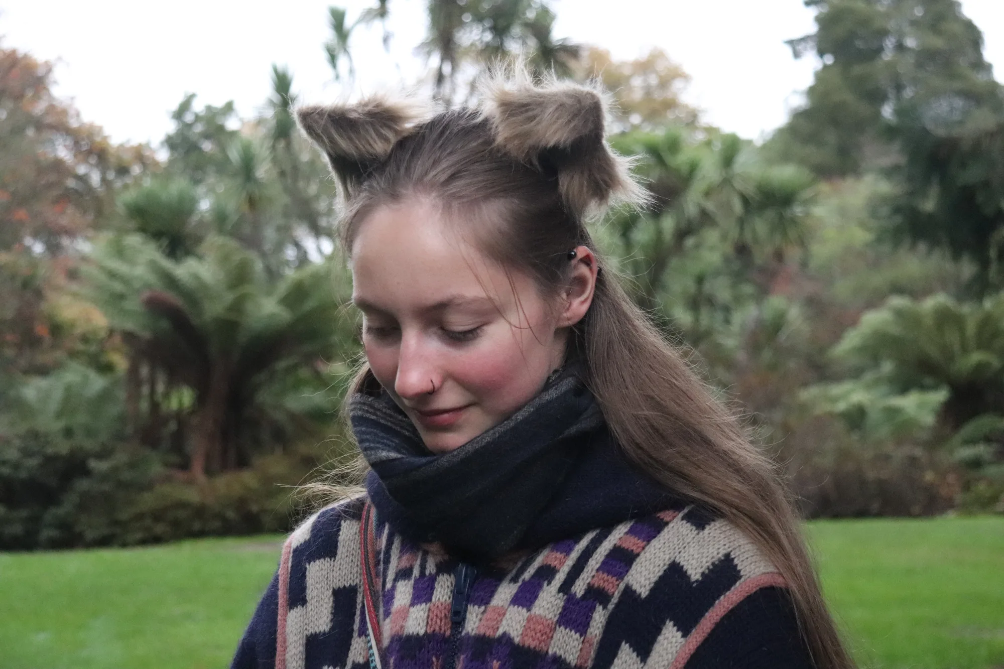 A woman with her hair styled into two pointed tufts looks down in a green garden