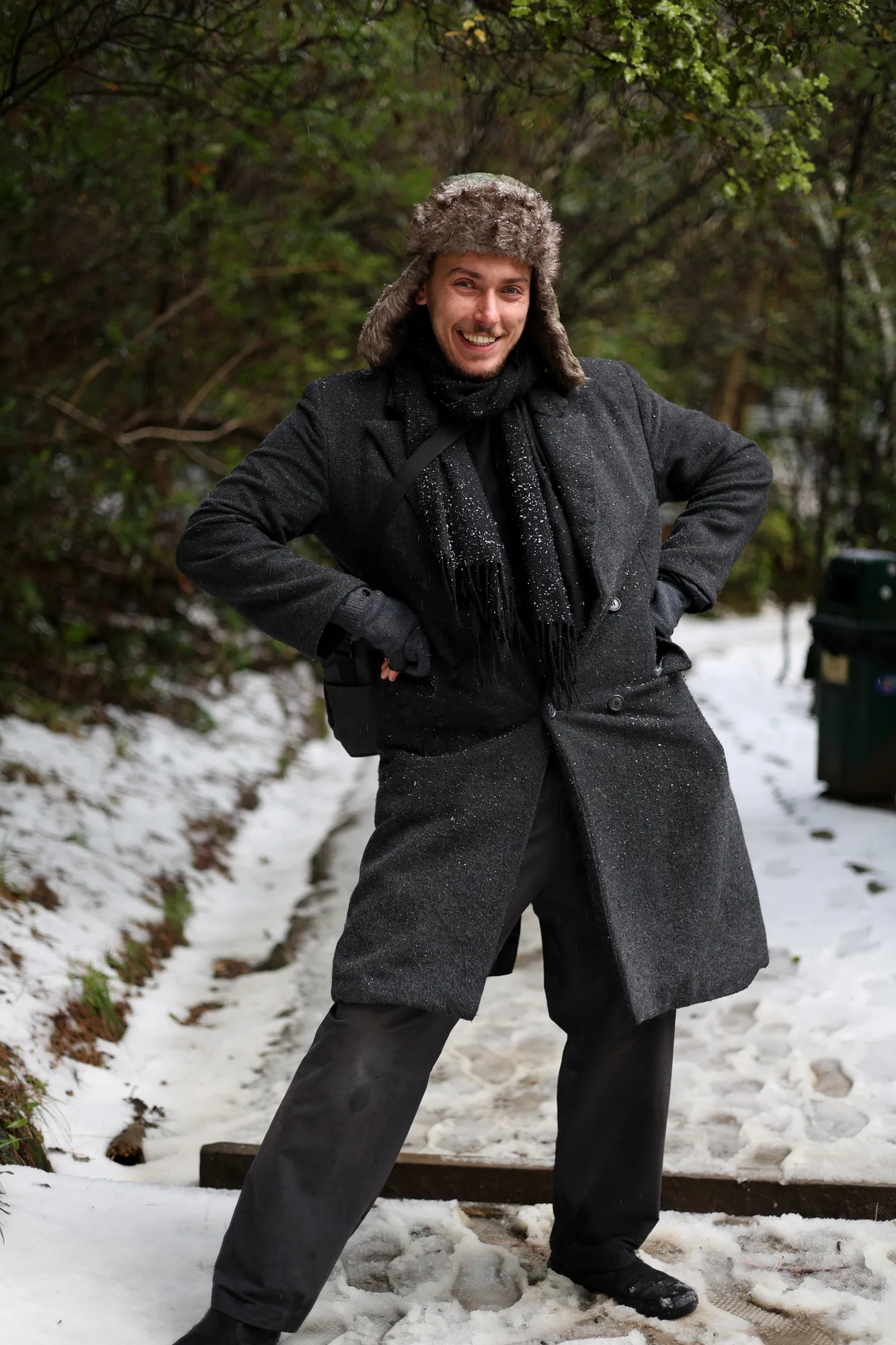 Kenan smiling in a dark coat and fur hat on a snowy forest path