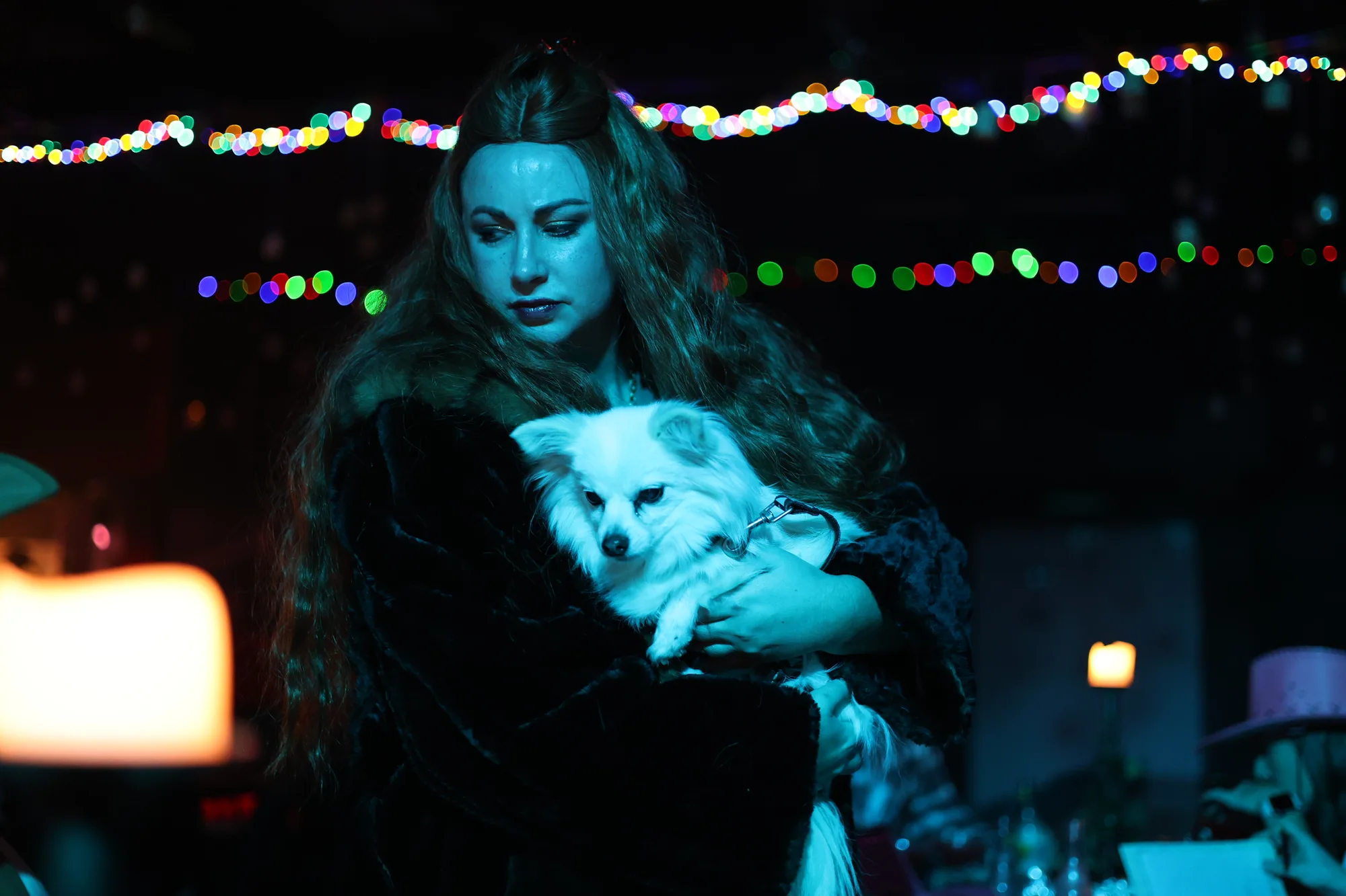 A woman in costume holds a small white dog beneath strings of coloured lights